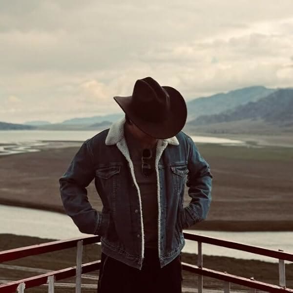 John Carrie in a denim jacket and wide-brimmed hat, looking down, with mountains and a lake behind him.
