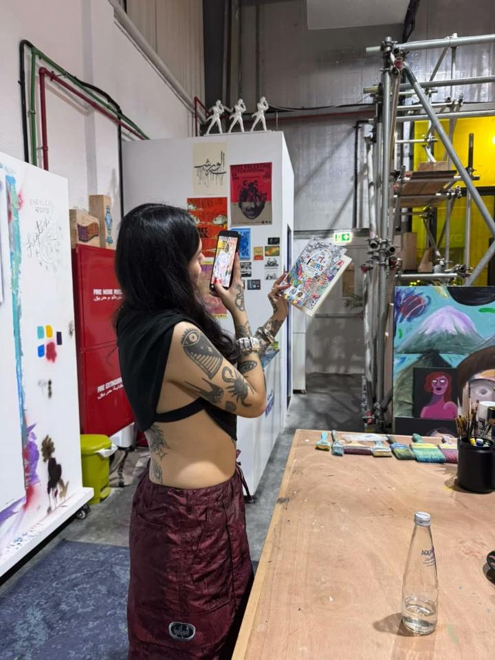 A woman in an art studio holds up the book and photographs its cover with her phone.