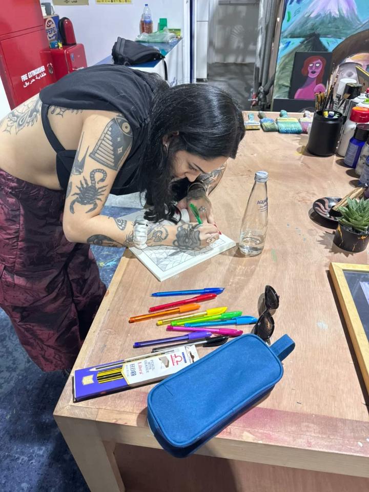 A woman with tattooed arms leans over a table in an art studio, hatching a page with a green pen.